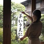 Japán nő tussal festett hiragana írásjegyeket mutat fel egy papíron, háttérben egy japánkert, japán tanulás